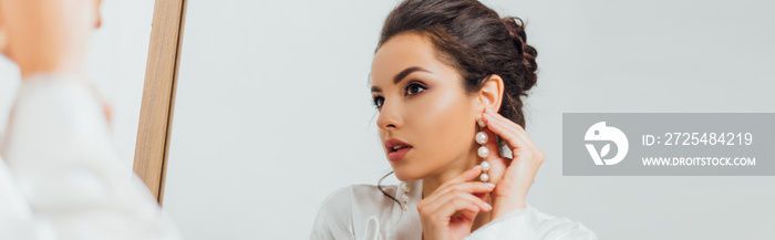 Panoramic shot of bride in silk robe putting on pearl earring near mirror