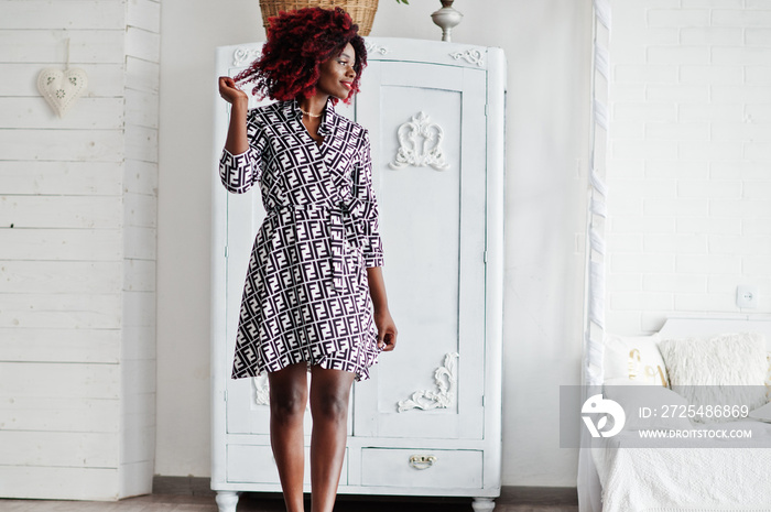 Fashionable tall african american model woman with red afro hair in dress posed at white room agains