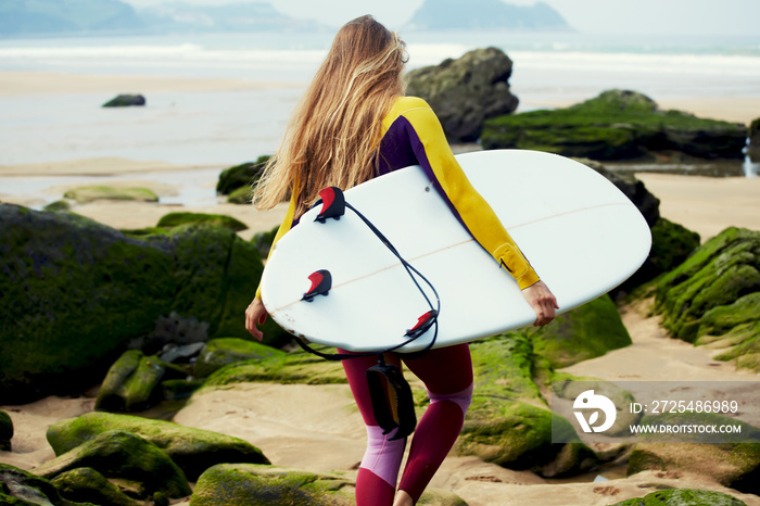 Blond long-haired girl dressed in wetsuit walking to the ocean ready to surfing, female surfer holdi