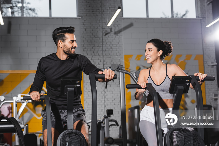 Smiling arabian sportsman looking at middle east sportswoman on elliptical trainer in gym.