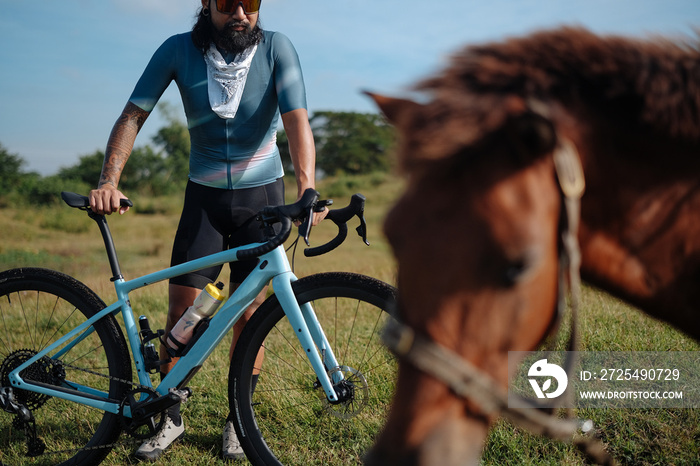 A young bearded cyclist is biking through a field with a horse