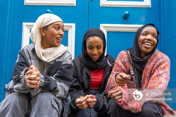 Three smiling women wearing�hijabs�sitting in front of blue door