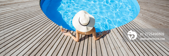 戴着大太阳帽，喝着鸡尾酒的女人在室外蓝水的圆形游泳池放松