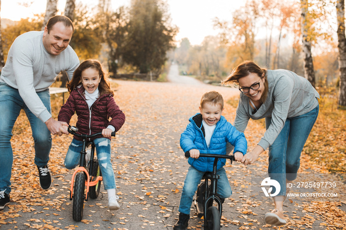 Young mother and father helping their kids to learn how to ride bicycles in the park against sunset.