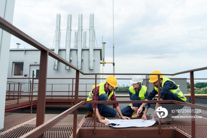 three engineers in helmets working with blueprints on roof together