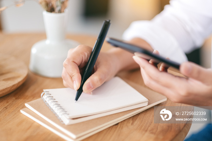 Closeup image of a woman using mobile phone while writing on a blank notebook on wooden table