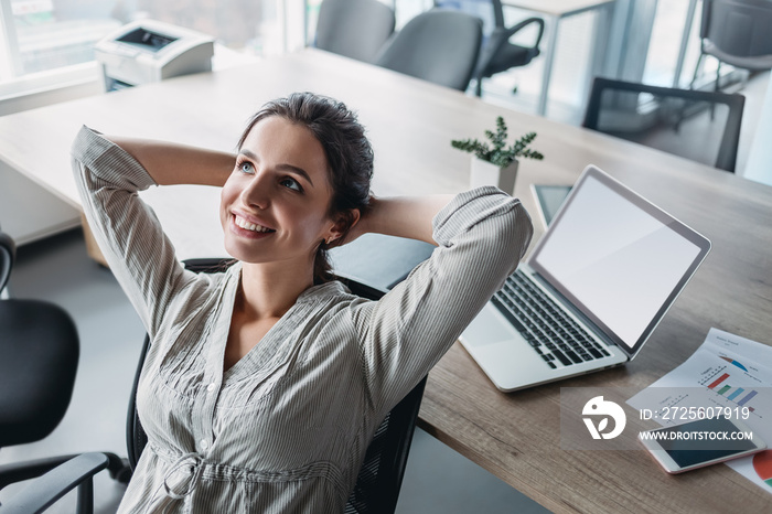 Happy businesswoman relaxing with hands behind head at office desk. Daydreaming concept