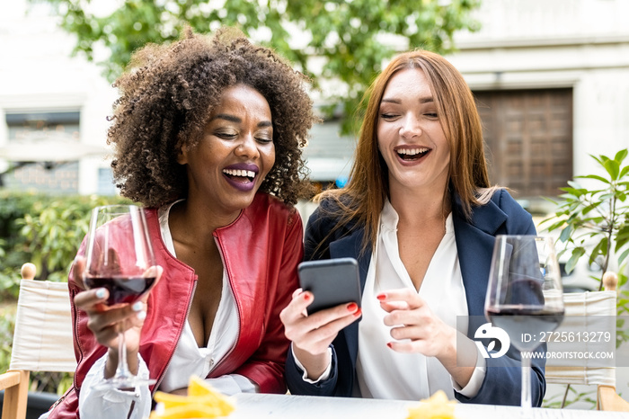 two adult woman having fun during happy hour after work, colleagues laughing while looking at smart 