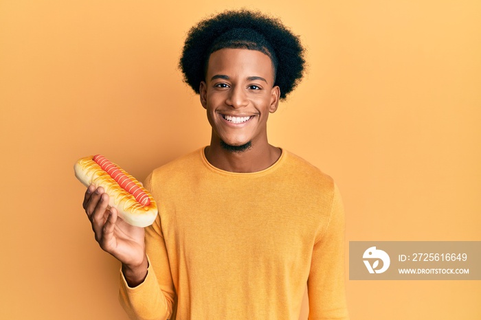 African american man with afro hair eating hotdog looking positive and happy standing and smiling wi