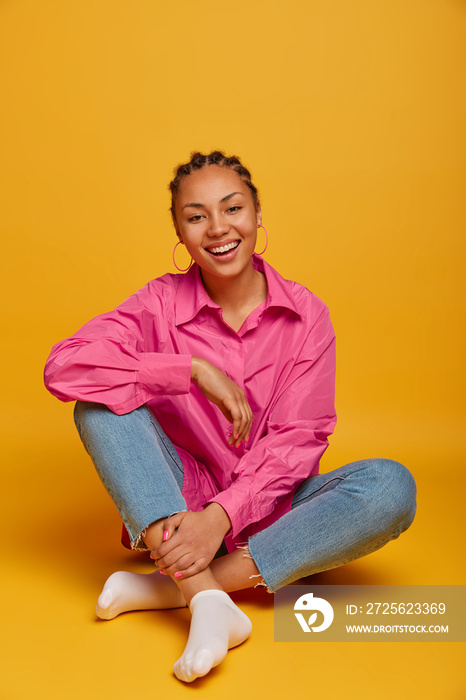 Vertical shot of good looking happy dark skinned teenage woman sits crossed legs on yellow floor, ha