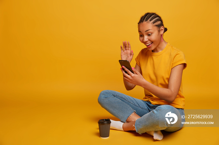 Horizontal view of ethnic millennial girl sits crossed legs on floor, enjoys browsing internet, has 