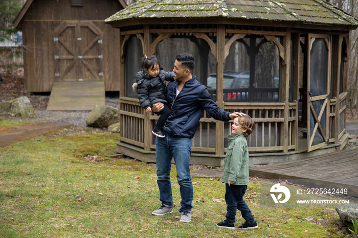 Father carrying cute daughter playing with boy while standing on grassy field against gazebo in fore