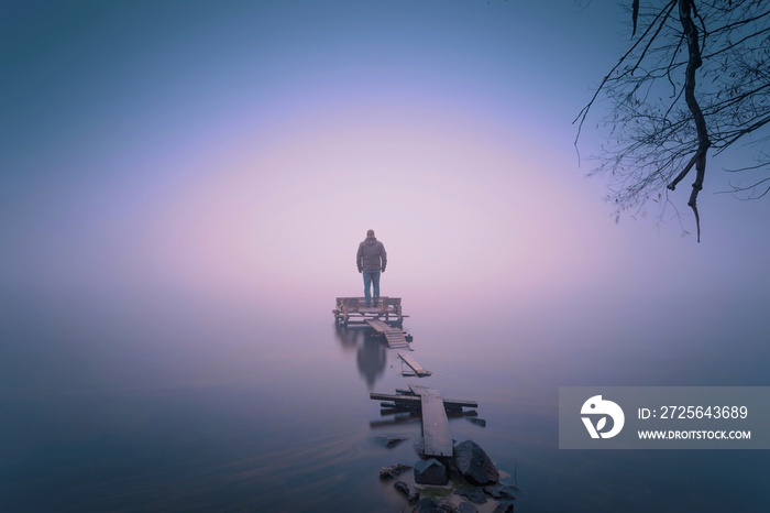 Lonely man, standing on the wooden jetty in the autumn foggy river with stones gangway.