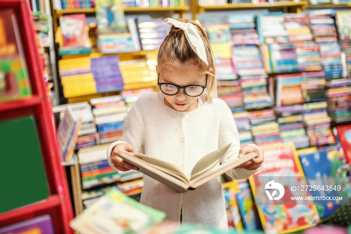 戴着眼镜的可爱好奇的高加索小女孩站在书店里读得很有趣
