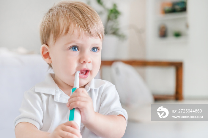 Small blue-eyed blond boy in a white polo shirt brushing his teeth with a toothbrush in the room