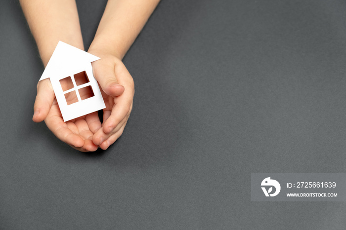 Top view on child hands holding white paper house on gray background. Family home and real estate co