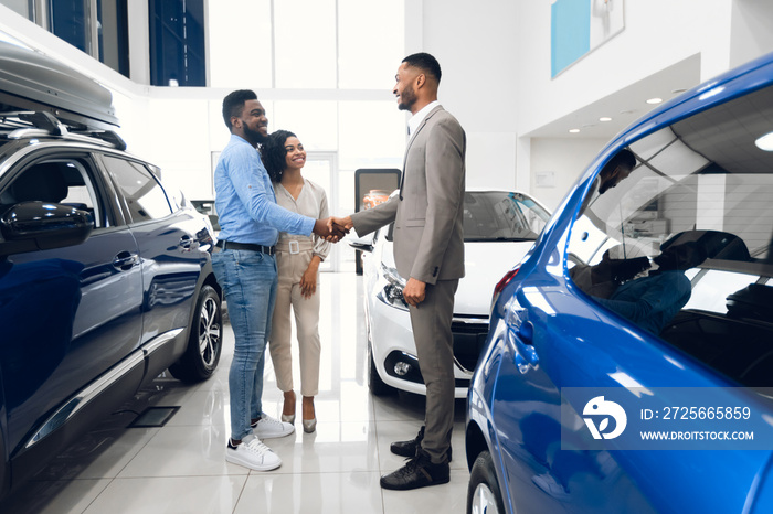 Happy Car Buyers Handshaking With Seller Standing In Dealership Showroom