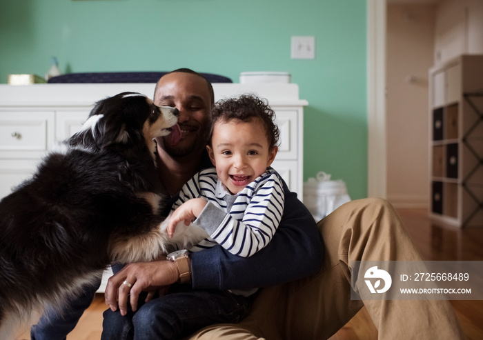 Dog licking man holding son while sitting against cabinet at home