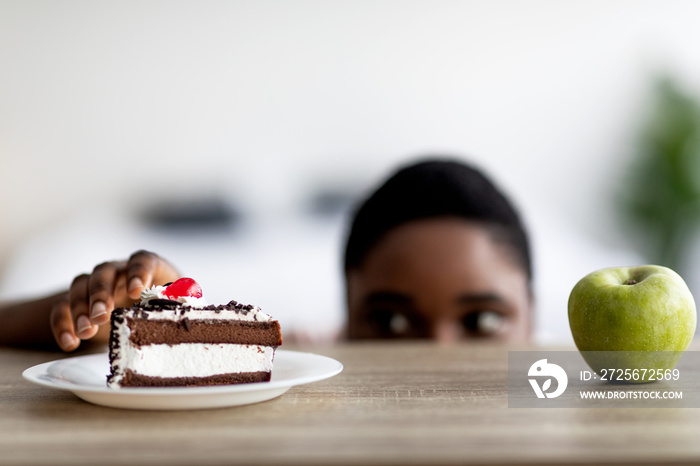 一位身材魁梧的非裔美国妇女在蛋糕和苹果之间做出选择，从桌子下面偷看食物，免费