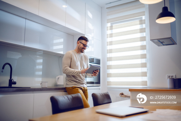 Positive beautiful Caucasian man dressed casual standing in modern kitchen and surfing on internet o