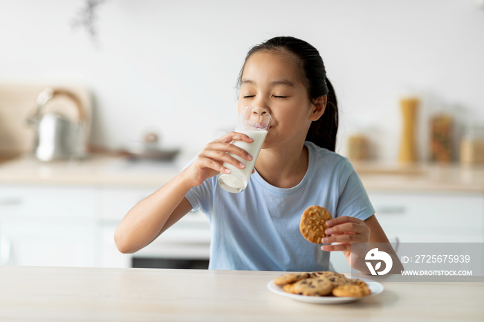 Asian girl drinking milk and enjoying cookies, sitting in kitchen and eating snack, free space