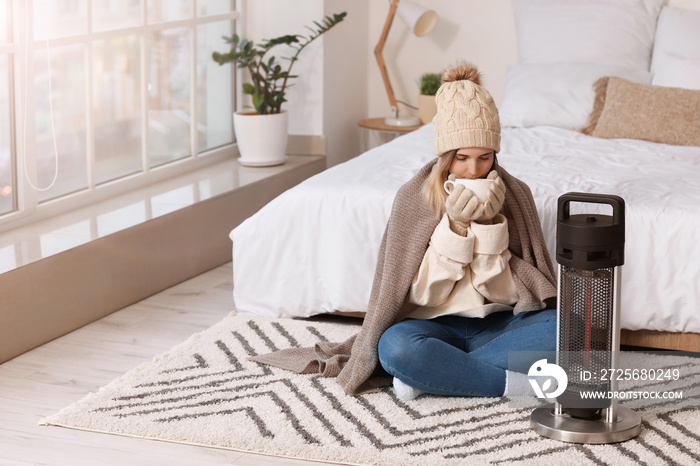 Young woman in warm clothes and with cup of tea sitting near electric heater at home. Concept of hea