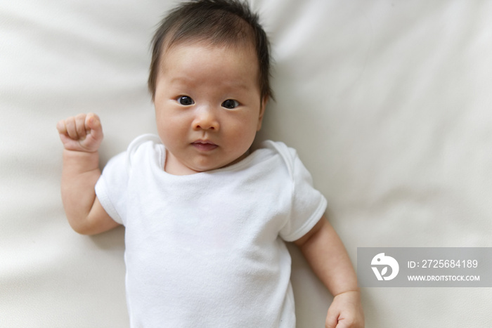 Asian newborn baby wearing white T-shirt lying on white sofa. Seen from top and close up at baby fac