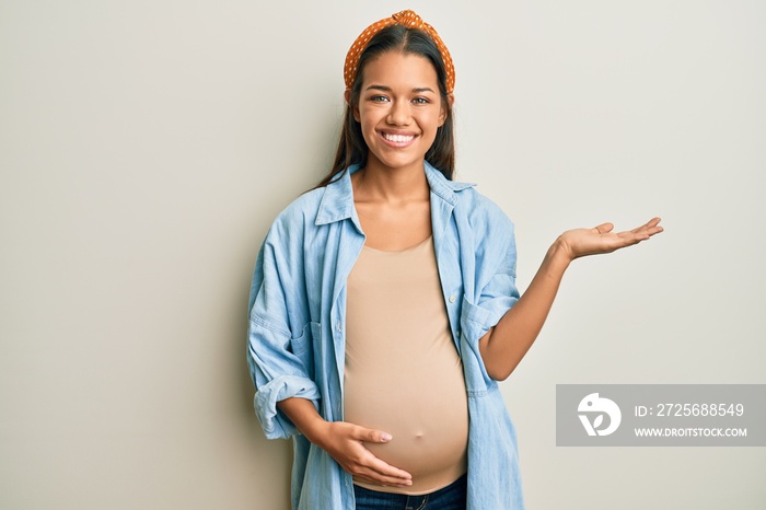 Beautiful hispanic woman expecting a baby, touching pregnant belly smiling cheerful presenting and p