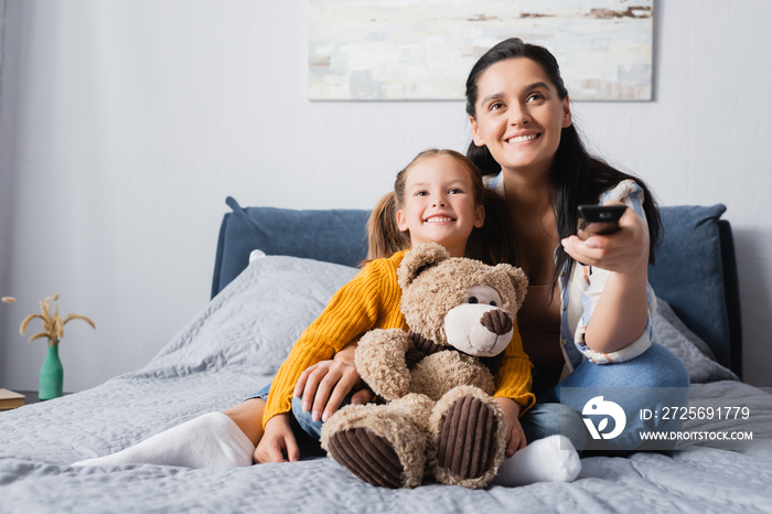 joyful woman holding tv remote controller while watching movie with daughter