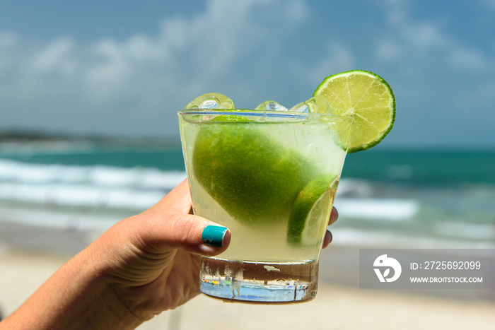 Drink caipirinha. Hand holding a caipirinha drink on a beach in Brazil with a blurred background.