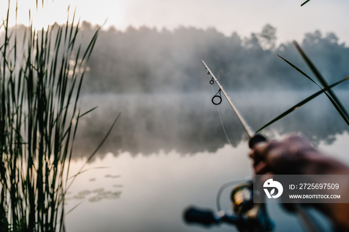 Fisherman with fishing rod, spinning reel on the background river bank. Sunrise. Fog against the bac