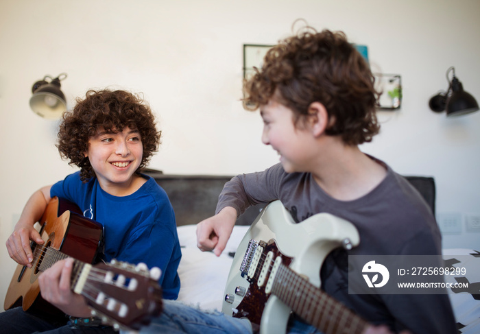 Smiling brothers practicing guitars while sitting on bed against wall at home