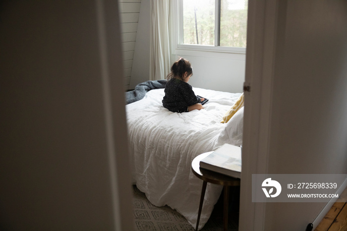 Rear view of girl using mobile phone while relaxing on bed at home seen through doorway