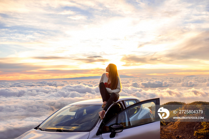身穿心形毛衣的年轻女子坐在车顶上欣赏美丽的云景