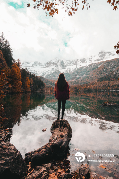 一名女性站在一块大石头上，看着美丽的湖泊和五颜六色的树木的垂直镜头