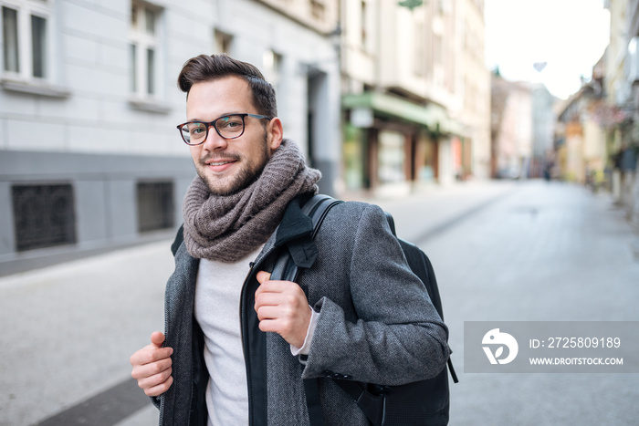 Portrait of a trendy young man with backpack walking in the city street. Looking at camera.