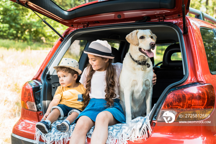 brother and sister sitting on car trunk with labrador dog