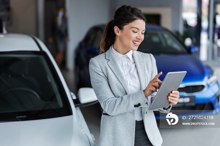 Smiling female seller in suit using tablet for looking which car is sold while standing in car salon