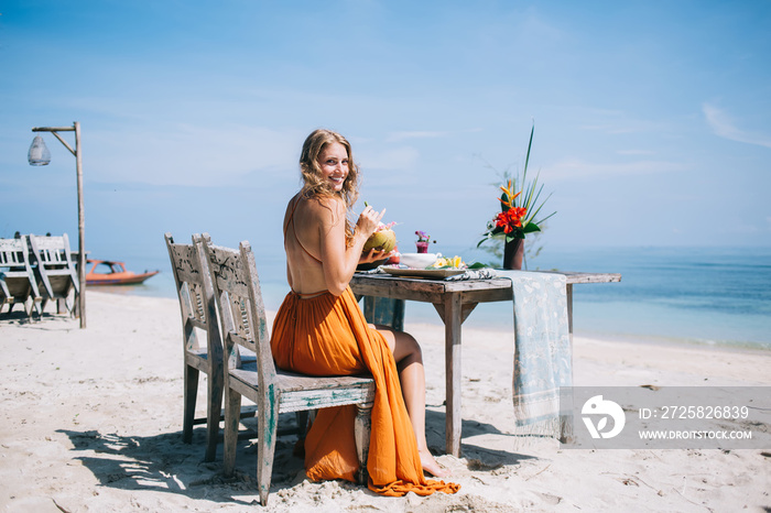 在清澈的蓝色沙滩上，坐在餐桌旁喝椰子汁的快乐成年女性