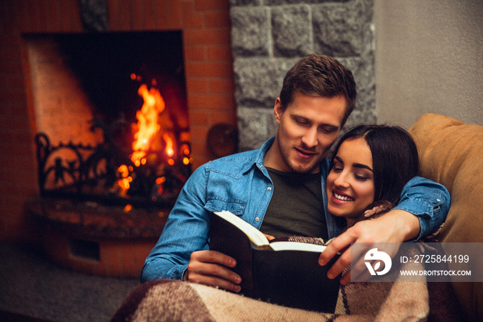 Positive and cheerful couple sit together. Young woman covered with blanket. Guy read book for her. 