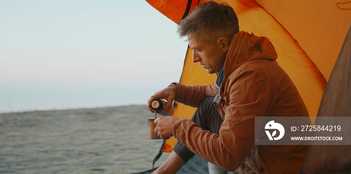 man traveler sitting at camp tent and pouring warm coffee from thermos in mug