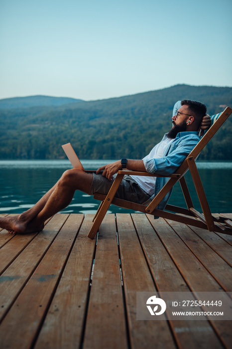 男性坐在湖边舒适的椅子上，一边用笔记本电脑，一边把它放在腿上。