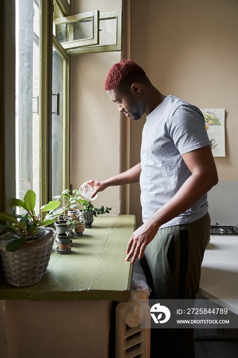 Man holding glass with water and watering green plants at the windowsill at home