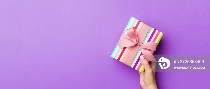 Flat lay of woman hands holding gift wrapped and decorated with bow on purple background with copy s