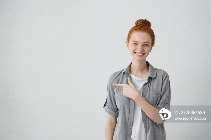 Smiling red-haired freckled woman in casual shirt posing against white studio wall pointing at copy 