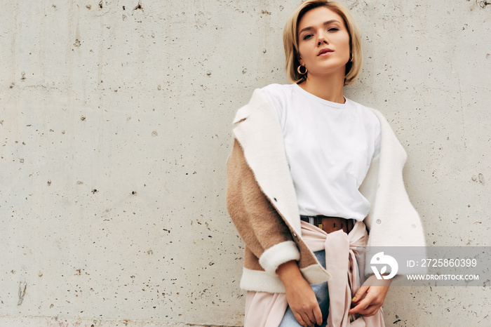 Image of stylish blonde young woman wearing white t-shirt and beige jacket, smiling and looking to t