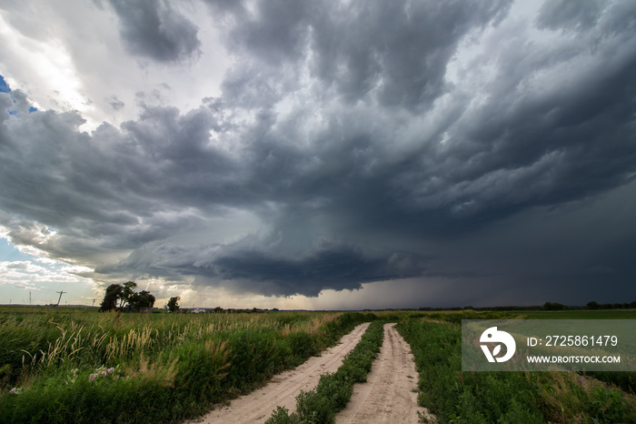 A supercell thunderstorm looms in the sky over a dirt road in farm country.