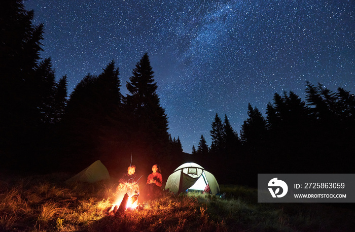情侣游客坐在两个帐篷附近明亮燃烧的篝火旁，享受bea的正视图