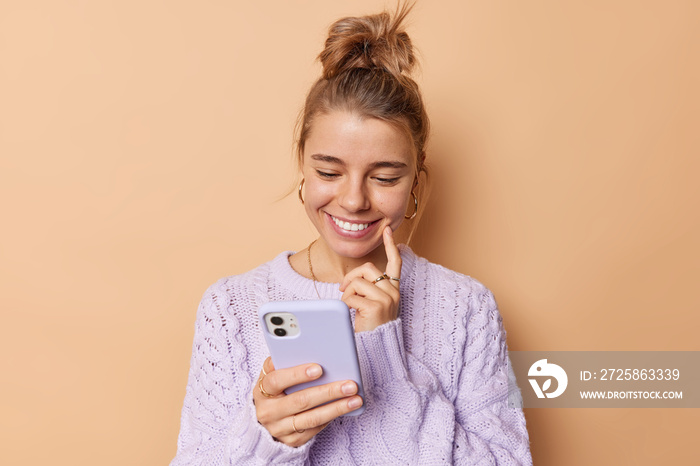Pleased young woman smiles happily keeps index finger on cheek concentrated into smartphone device r