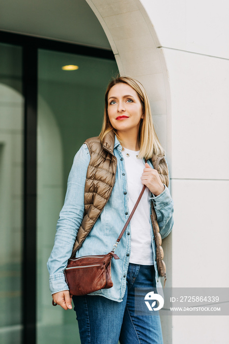Portrait of beautiful young woman posing outdoors, wearing denim clothes, brown down jacket and cros